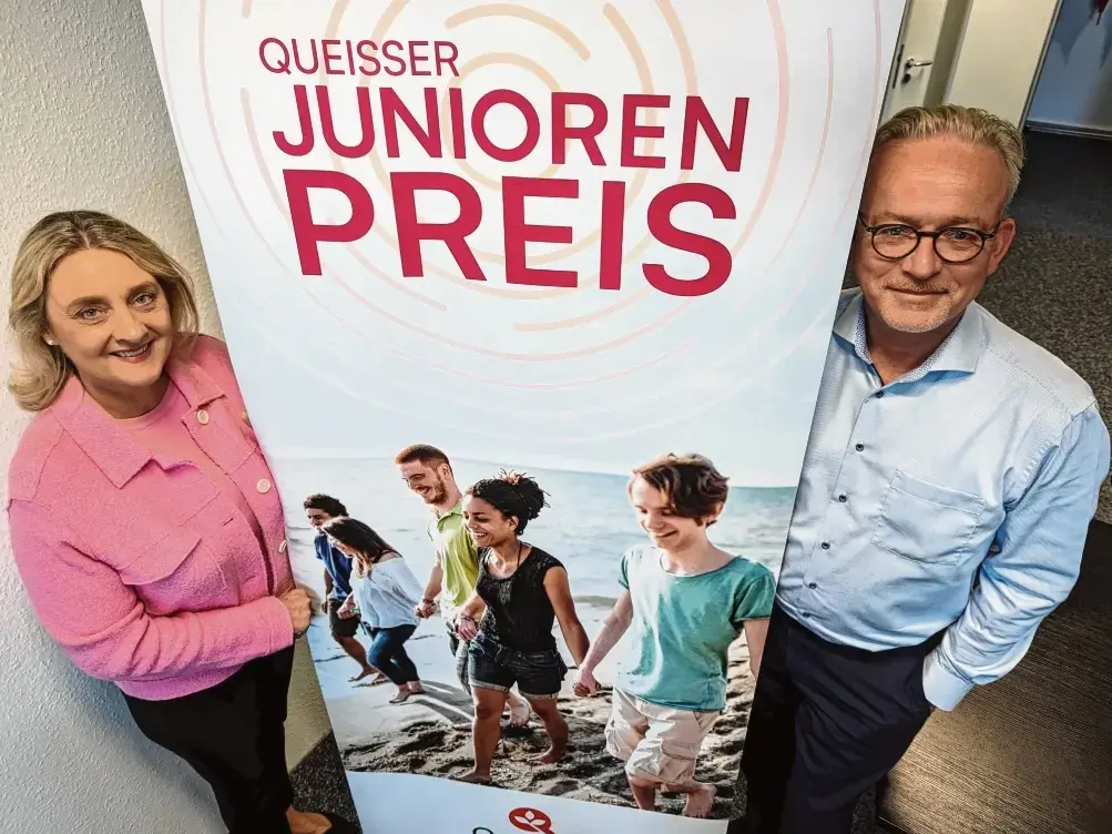 Zwei Personen stehen links und rechts neben einem Roll-up mit der Aufschrift ‚Queisser Junioren Preis‘ und einem Foto von Kindern, die am Strand entlanglaufen.