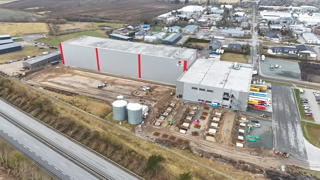 Luftaufnahme des Queisser-Logistikzentrums mit angebauter Lagerhalle, drei großen Silos, Baustellenbereich mit Fundamenten und angrenzender Bundesstraße.