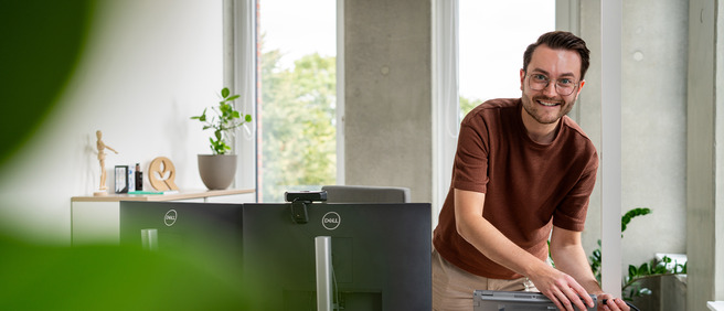 Junger Mann mit Brille lächelt im modernen Büro an seinem Arbeitsplatz.