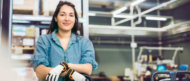 Junge Frau mit Arbeitshandschuhen steht in einer modernen Produktionshalle.