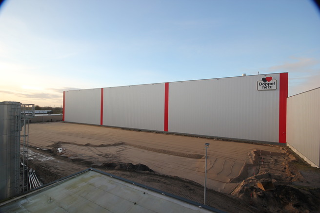 Vorbereitete Baufläche vor der Fassade des Queisser-Logistikzentrums mit rotem Doppelherz-Logo, planiertes Sandbett für nachfolgende Baumaßnahmen