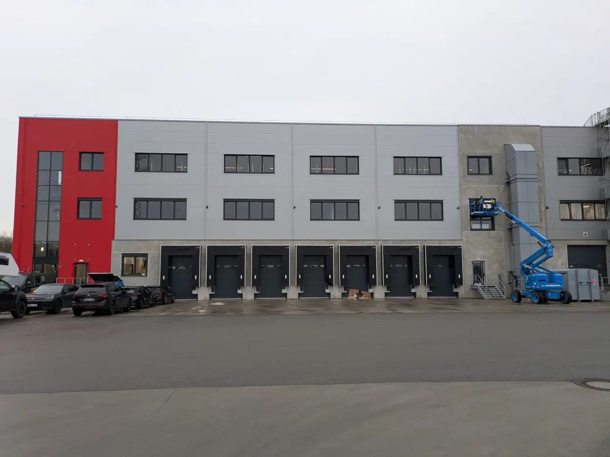 Das Bild zeigt ein modernes Lagergebäude mit einer roten und grauen Fassade. Mehrere Ladetüren sind sichtbar, und ein blauer Arbeitslift steht auf dem Platz. Der Himmel ist bedeckt, und die Umgebung wirkt industriell.