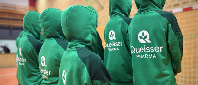 Sportteam mit grünen Kapuzenpullovern, auf dem Rücken das Queisser-Logo, von hinten fotografiert in einer Sporthalle.