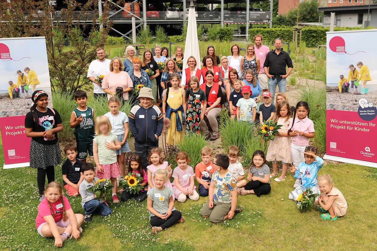 Eine große Gruppe von Erwachsenen und Kindern steht in einem Garten vor zwei Plakaten, die ein Projekt für Kinder unterstützen. Einige Kinder halten Blumen, während andere lächeln oder in die Kamera schauen. Die Szene vermittelt eine Atmosphäre von Gemeinschaft und Freude.