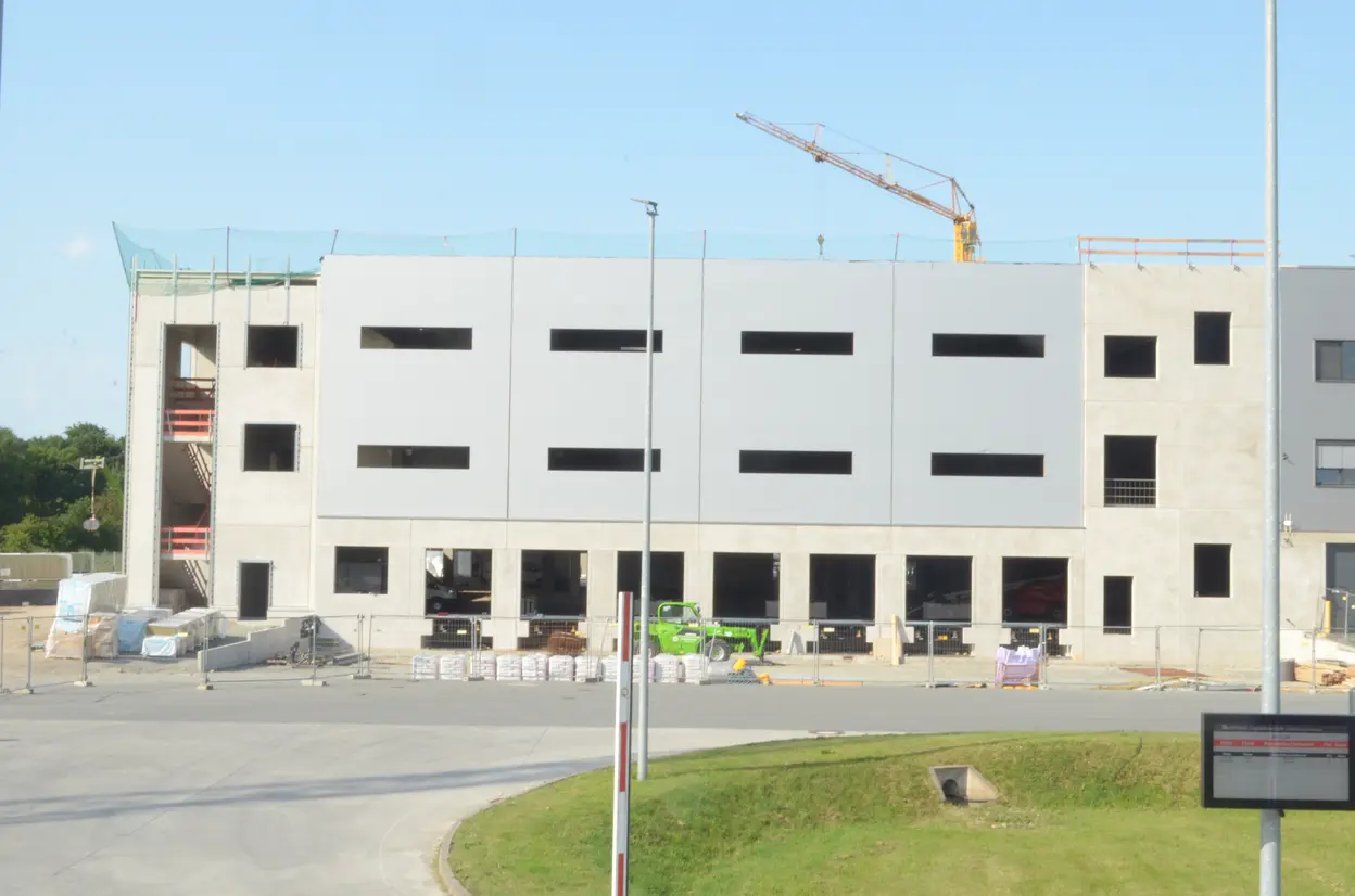 Eine Baustelle eines Gebäudes ist zu sehen, das aus grauen Betonwänden besteht. Auf mehreren Etagen sind noch Fenster und Türen nicht eingebaut. Ein Kran hebt Materialien an, und verschiedene Baustellenmaterialien befinden sich am Boden. Eine asphaltierte Fläche führt zur Baustelle.