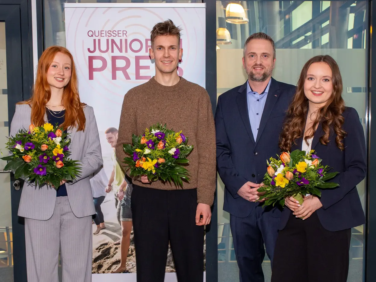 Vier Personen stehen vor einem Banner mit der Aufschrift "Queisser Junior Preis". Alle halten bunte Blumensträuße in der Hand. Die Gruppe besteht aus zwei Frauen und zwei Männern, die in formeller Kleidung gekleidet sind. Die Atmosphäre wirkt feierlich und freundlich.