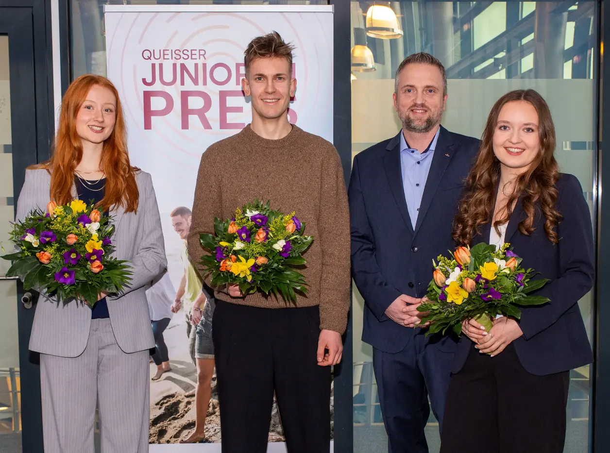 Vier Personen stehen vor einem Banner mit der Aufschrift "Queisser Junior Preis". Alle halten bunte Blumensträuße in der Hand. Die Gruppe besteht aus zwei Frauen und zwei Männern, die in formeller Kleidung gekleidet sind. Die Atmosphäre wirkt feierlich und freundlich.