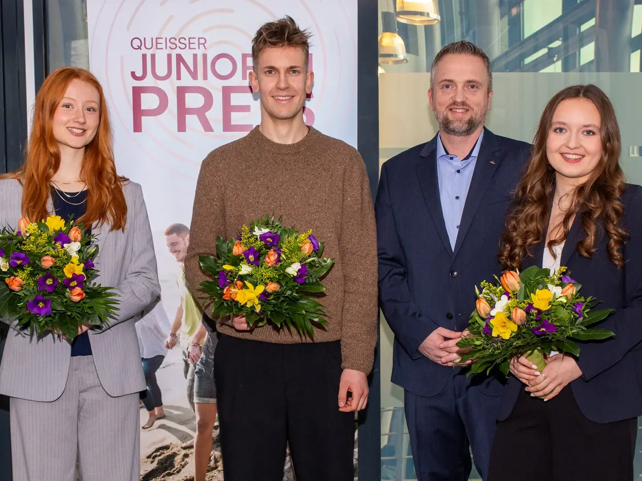 Vier Personen stehen vor einem Banner mit der Aufschrift "Queisser Junior Preis". Alle halten bunte Blumensträuße in der Hand. Die Gruppe besteht aus zwei Frauen und zwei Männern, die in formeller Kleidung gekleidet sind. Die Atmosphäre wirkt feierlich und freundlich.