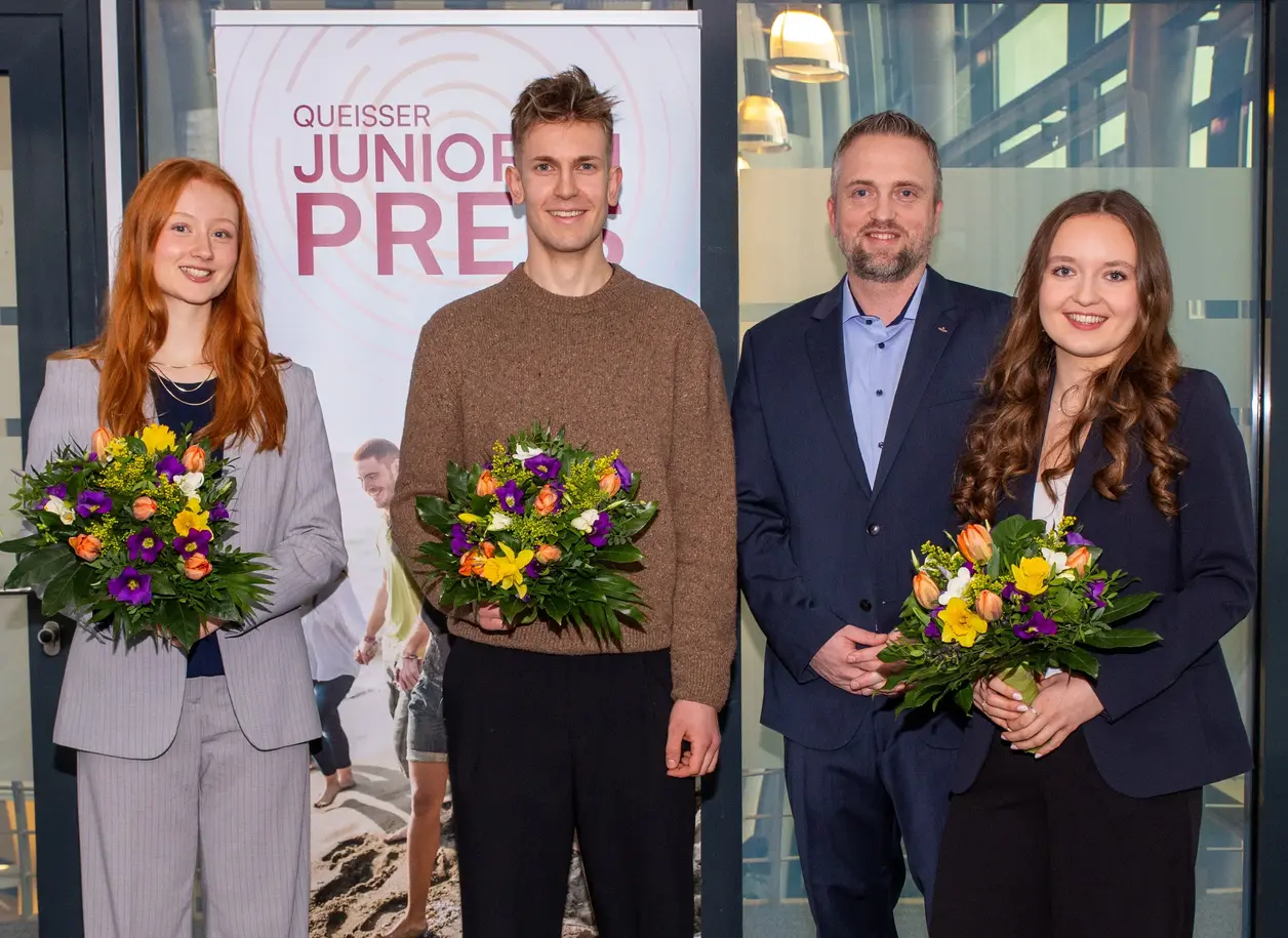 Vier Personen stehen vor einem Banner mit der Aufschrift "Queisser Junior Preis". Alle halten bunte Blumensträuße in der Hand. Die Gruppe besteht aus zwei Frauen und zwei Männern, die in formeller Kleidung gekleidet sind. Die Atmosphäre wirkt feierlich und freundlich.