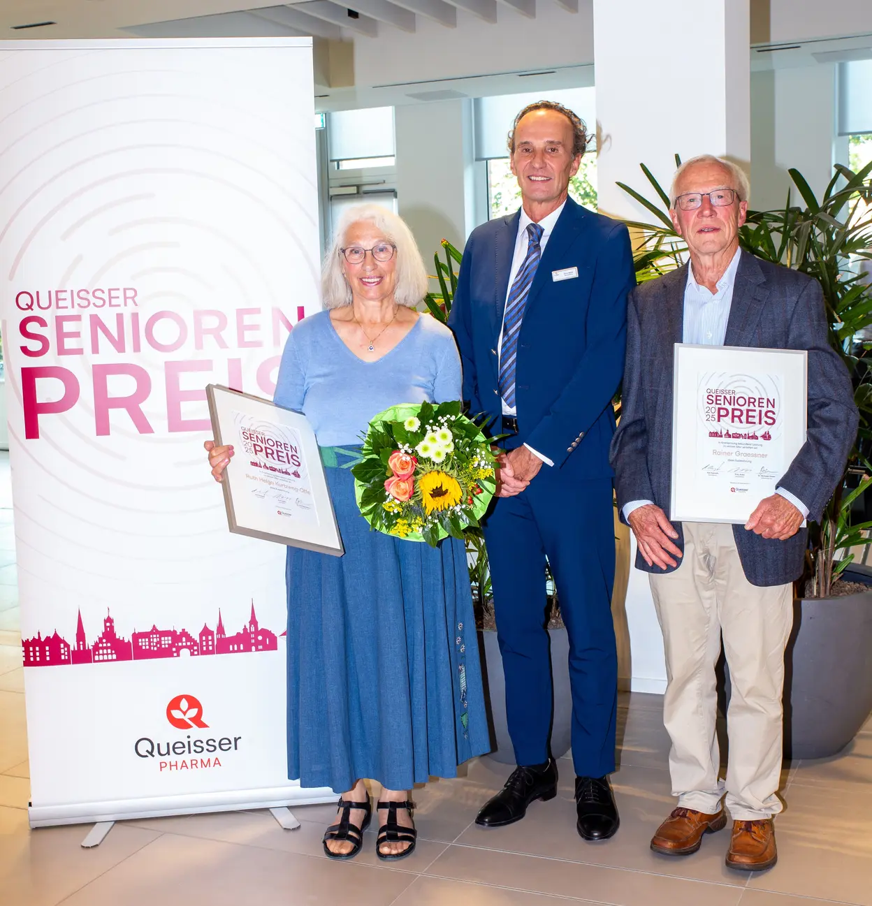 Drei Personen stehen vor einem Banner mit der Aufschrift "Queisser Senioren Preis". Eine Frau hält einen Blumenstrauß und eine Urkunde, während ein Mann in der Mitte mit einem Anzug ein Lächeln zeigt. Der dritte Mann hält ebenfalls eine Urkunde.