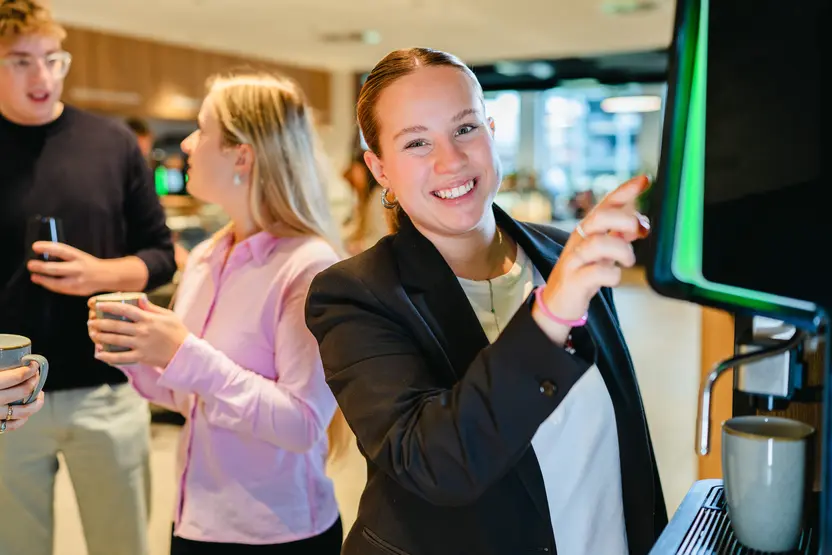 Eine junge Frau in einem schwarzen Blazer lächelt, während sie an einem Getränkespender steht. Sie zeigt mit dem Finger auf das Gerät. Zwei weitere Personen, ein Mann und eine Frau, stehen im Hintergrund und halten Tassen in der Hand.