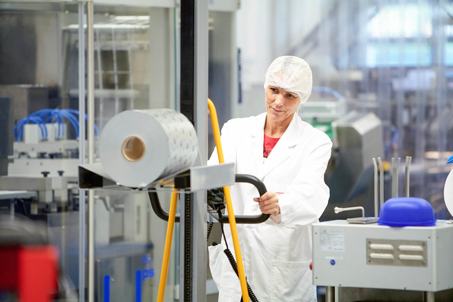 Mitarbeiterin in weißem Laborkittel und Haarnetz bedient einen Hubwagen neben einer Verpackungsmaschine mit großer Kunststoff-Folienrolle.