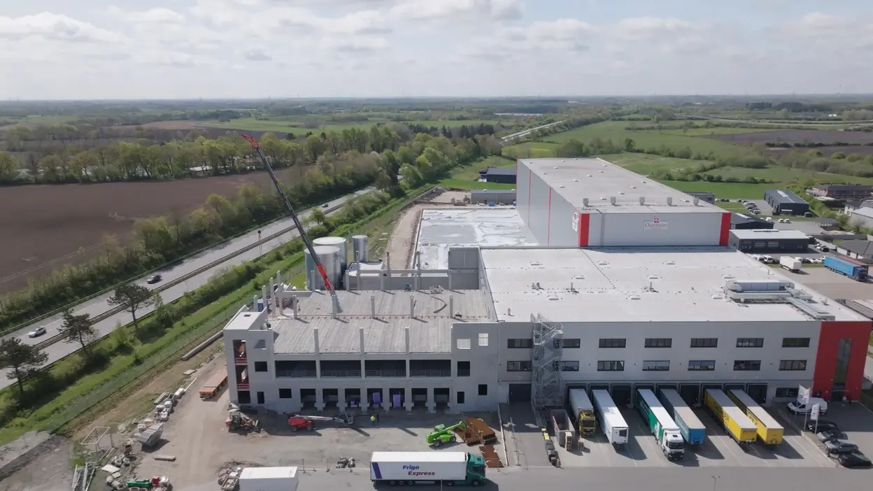 Aerial view of a large industrial building under construction, with loading docks and trucks parked in front. Nearby, cranes and construction equipment are visible. Surrounding areas include green fields and a road, with a few vehicles visible on the road. The scene conveys an active industrial setting.