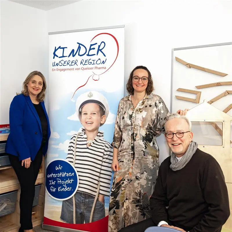 Mitarbeiter*innen von Queisser Pharma vor einem Roll-Up, das ein Kind in Matrosenverkleidung mit dem Logo "Kinder unserer Region" abbildet